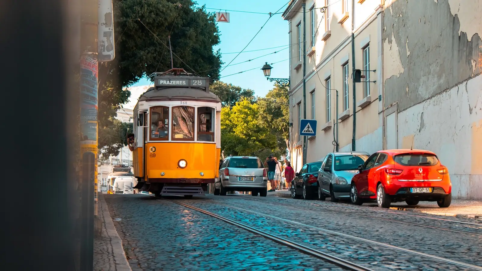 A yellow tram 28 to Prazeres on a sunlit cobbled street in Lisbon